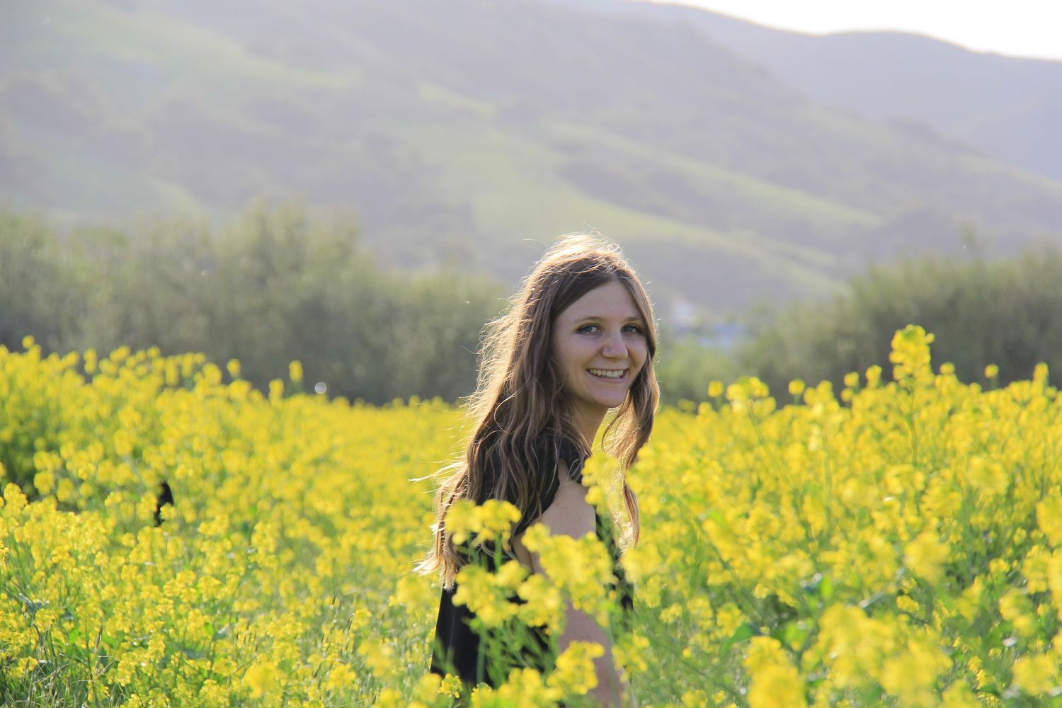 Person smiling among vibrant yellow wildflowers with rolling hills in background.