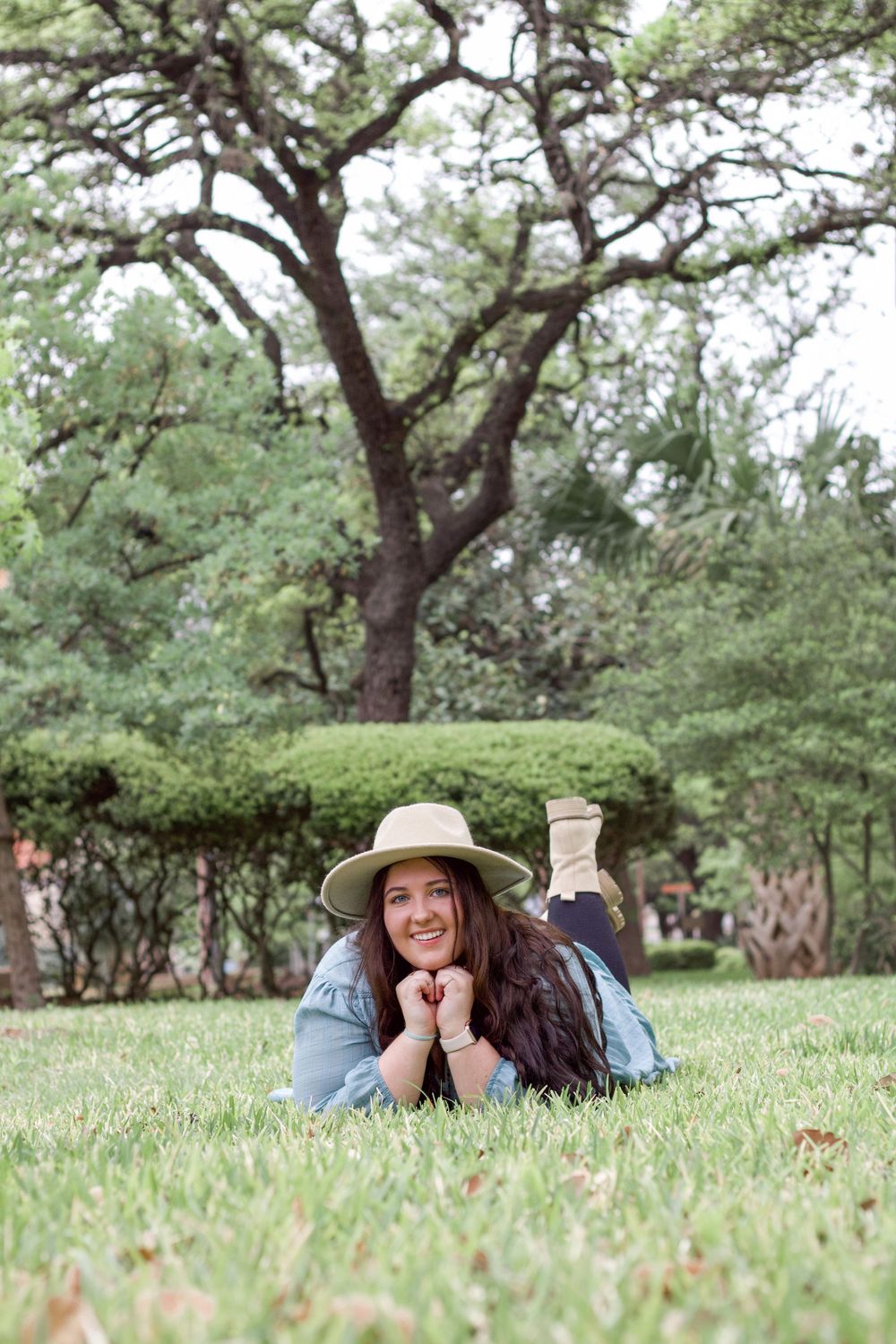 Relaxed outdoor portrait of someone lying on grass wearing a sun hat under a large oak tree.