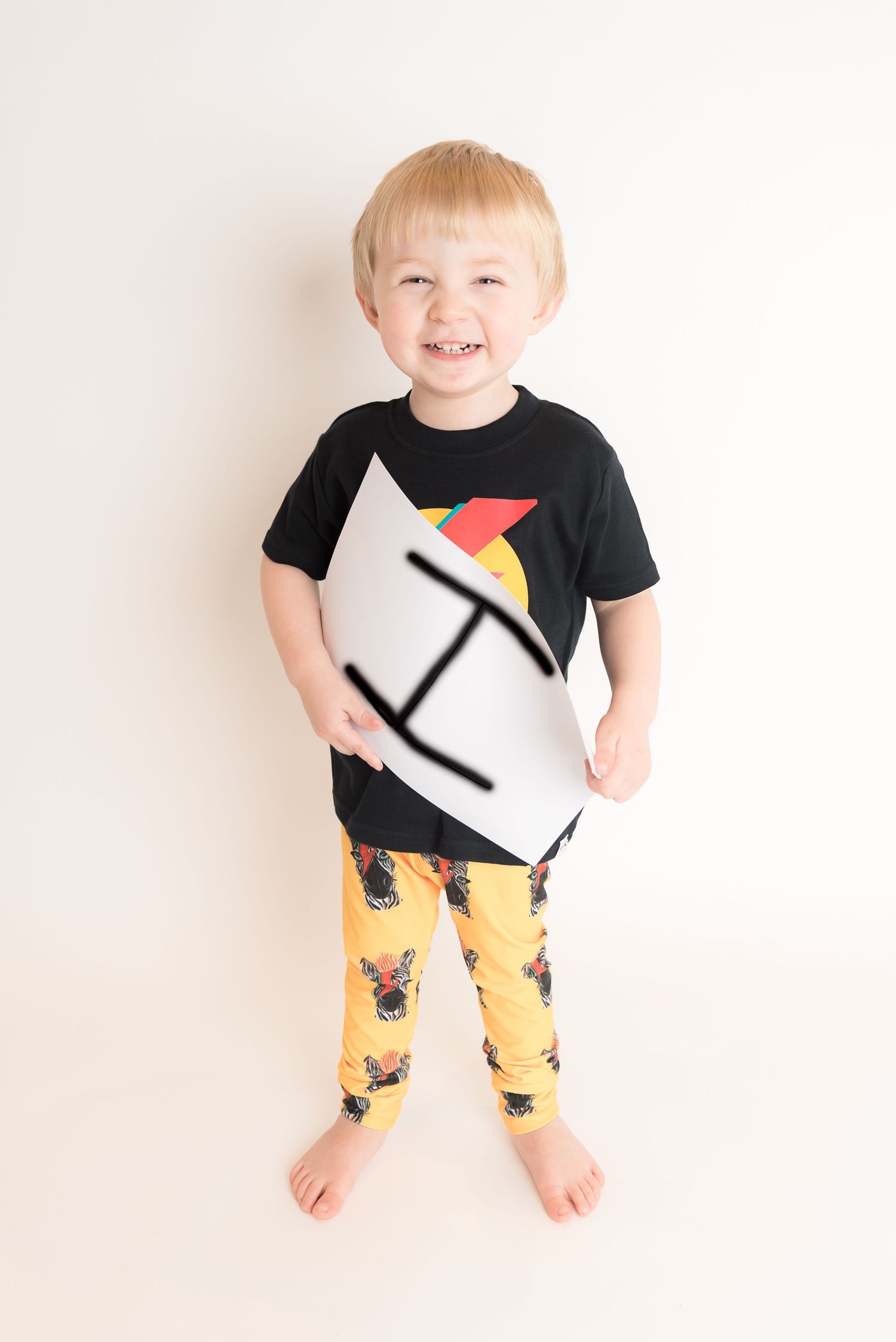 Young person in black geometric pattern shirt and yellow pants standing against white wall.