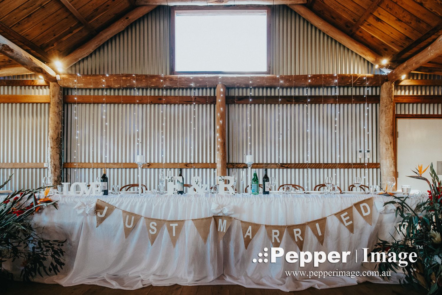 Rustic barn wedding head table decorated with a Just Married banner, tropical florals, and warm overhead lighting.