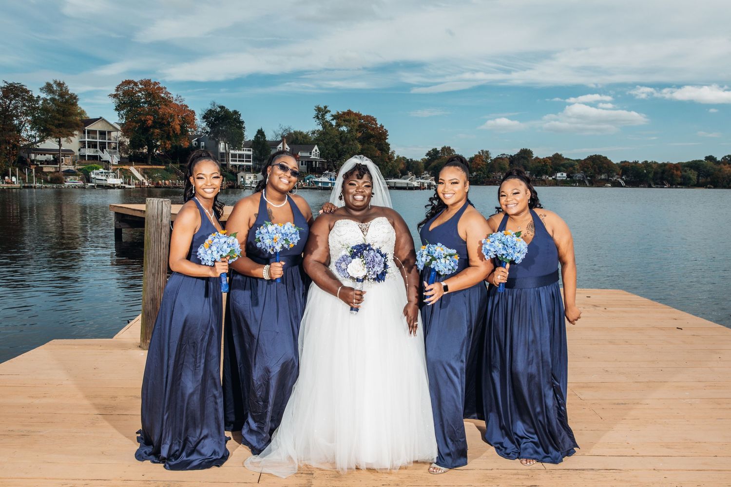 Bride in white gown poses with four bridesmaids in navy blue dresses holding blue bouquets on a waterfront dock.