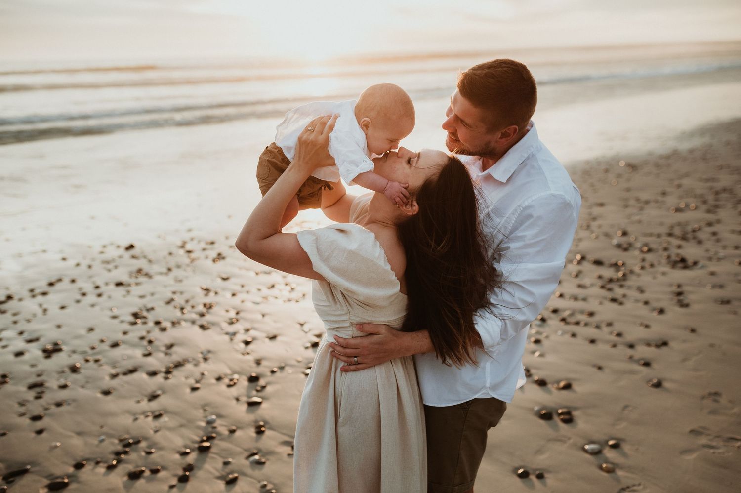 A joyful couple plays with their baby on a sandy beach during sunset.