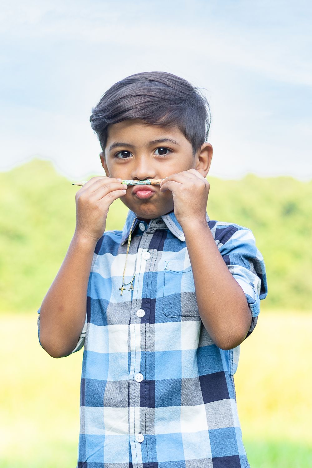 Student in blue plaid shirt makes playful mustache gesture with hair outdoors.