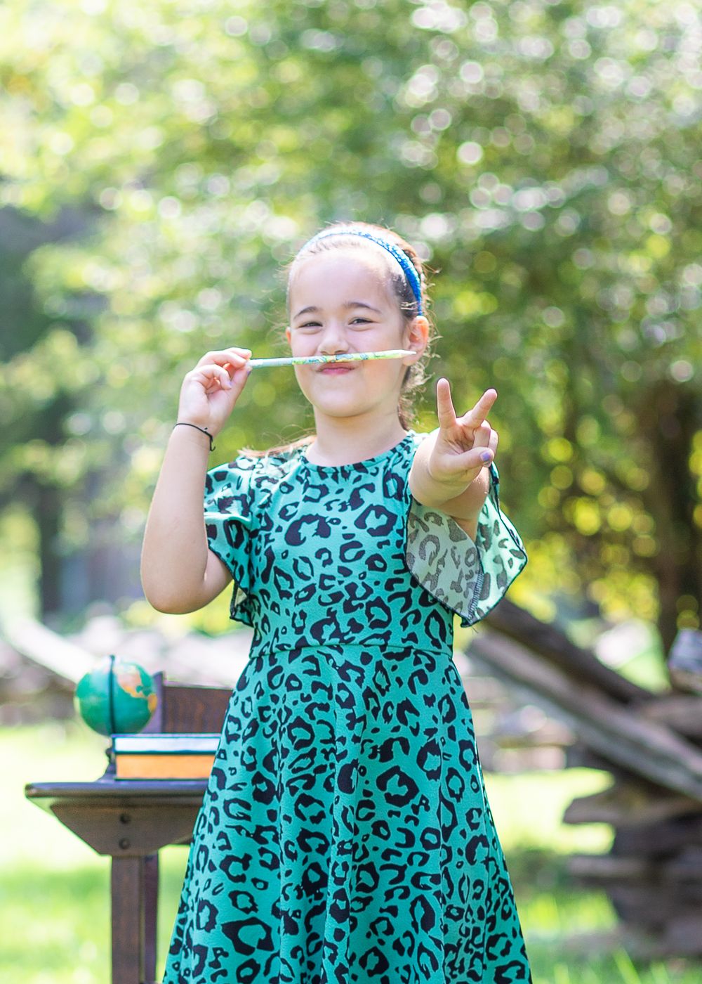 Student in teal patterned dress makes peace sign while creating hair mustache outdoors.