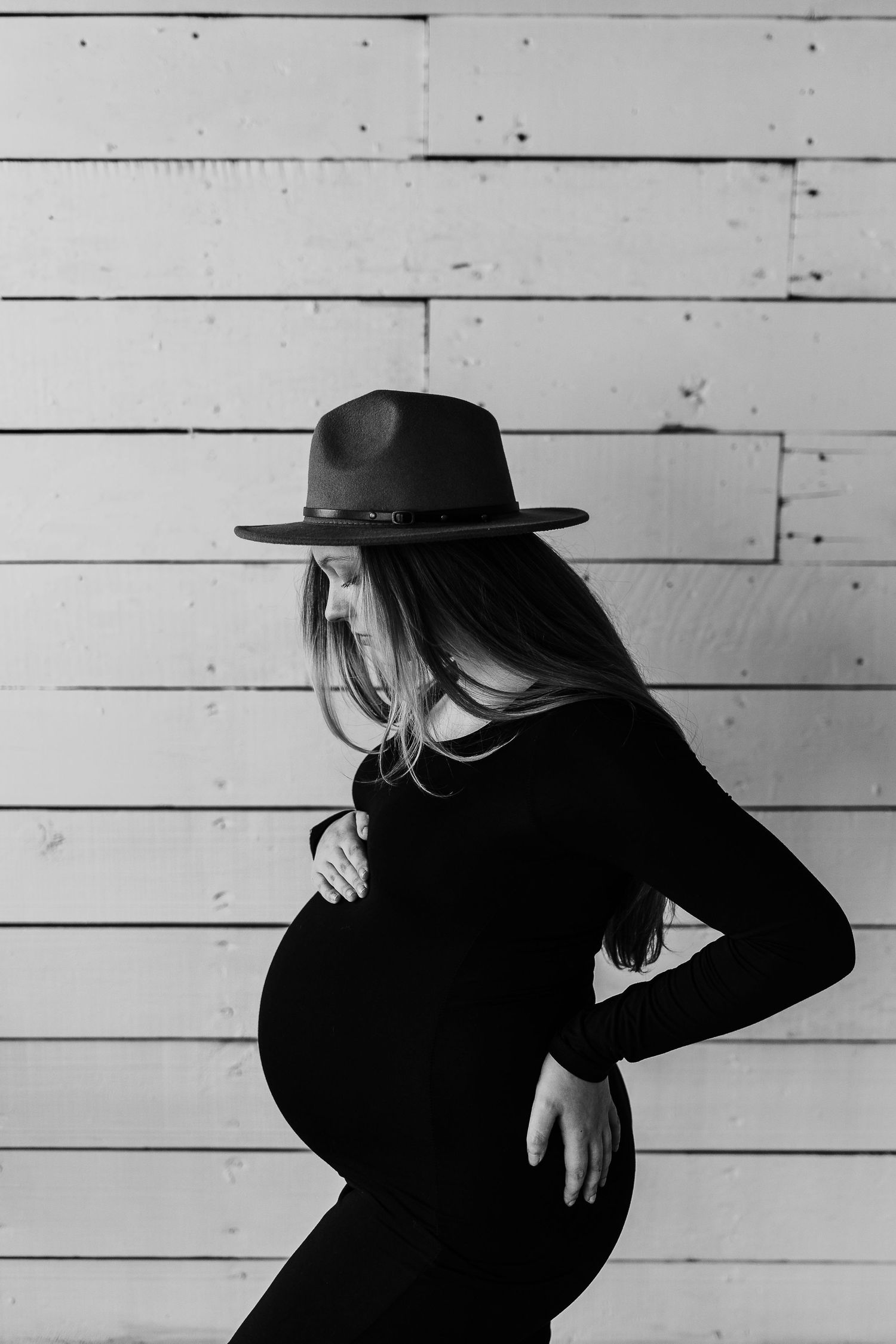 Silhouette of pregnant woman in black dress and hat against white wooden wall.