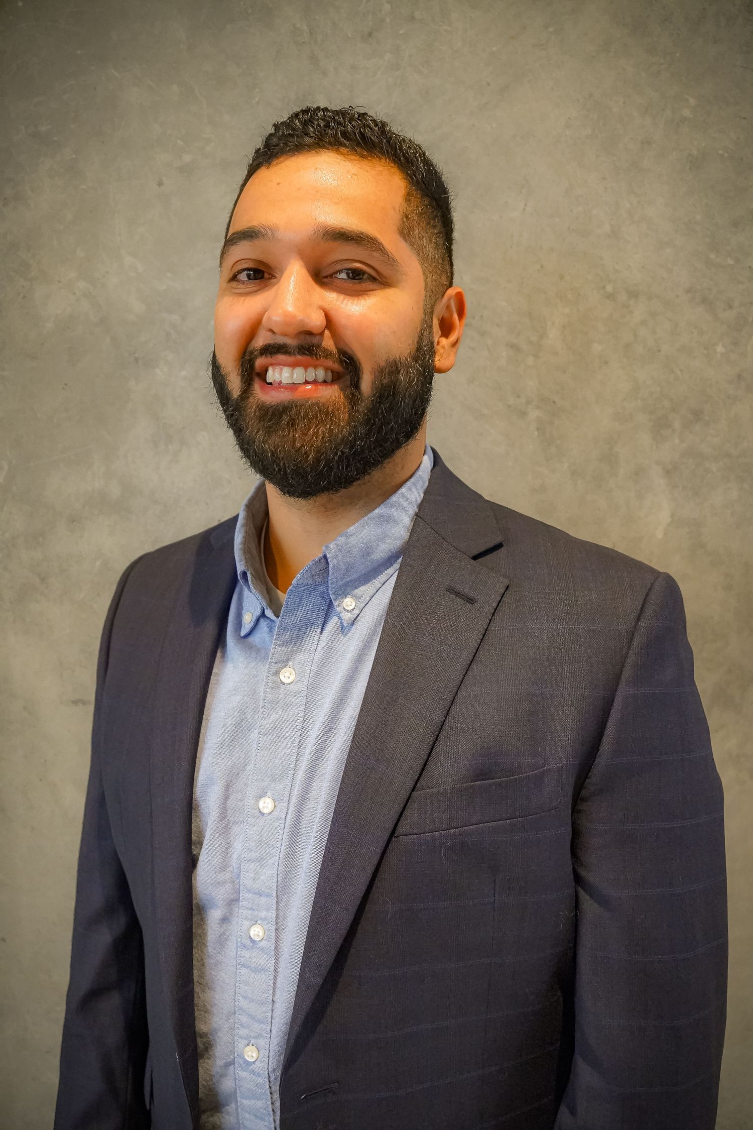 Professional headshot of bearded person in business attire against textured wall.