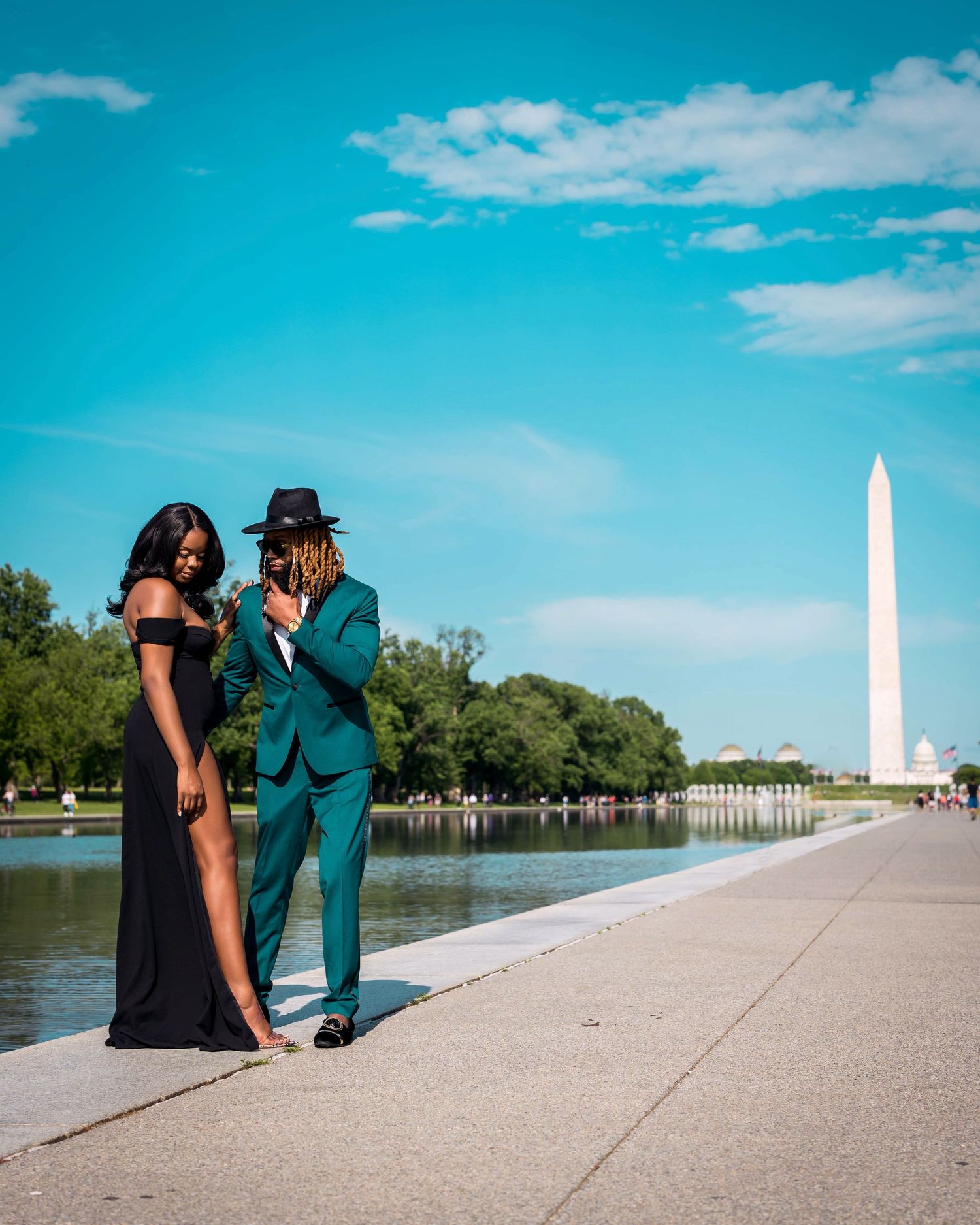 Stylish couple in formal wear poses near reflecting pool with Washington Monument visible.