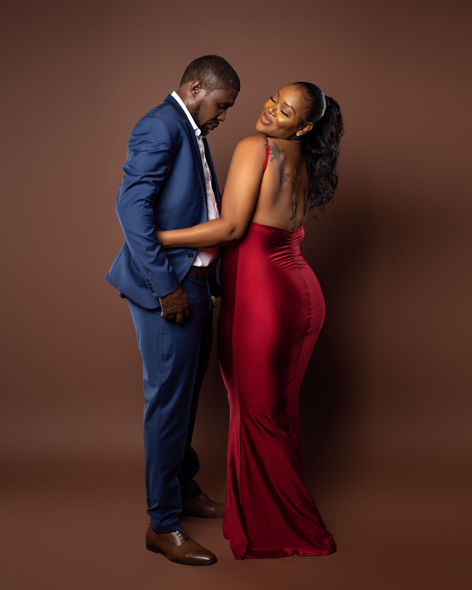 Couple in formal attire shares romantic moment against brown background.