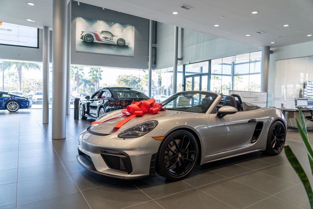 Silver Porsche GT4 sports car with red bow displayed in modern dealership showroom.