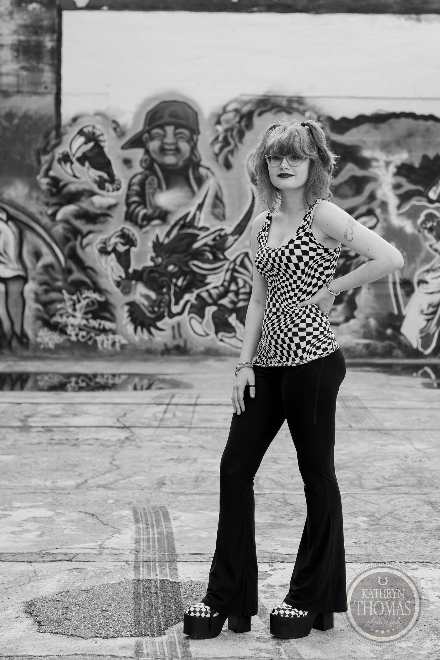 A black and white photo of someone in retro style against graffiti backdrop wearing checkered top.