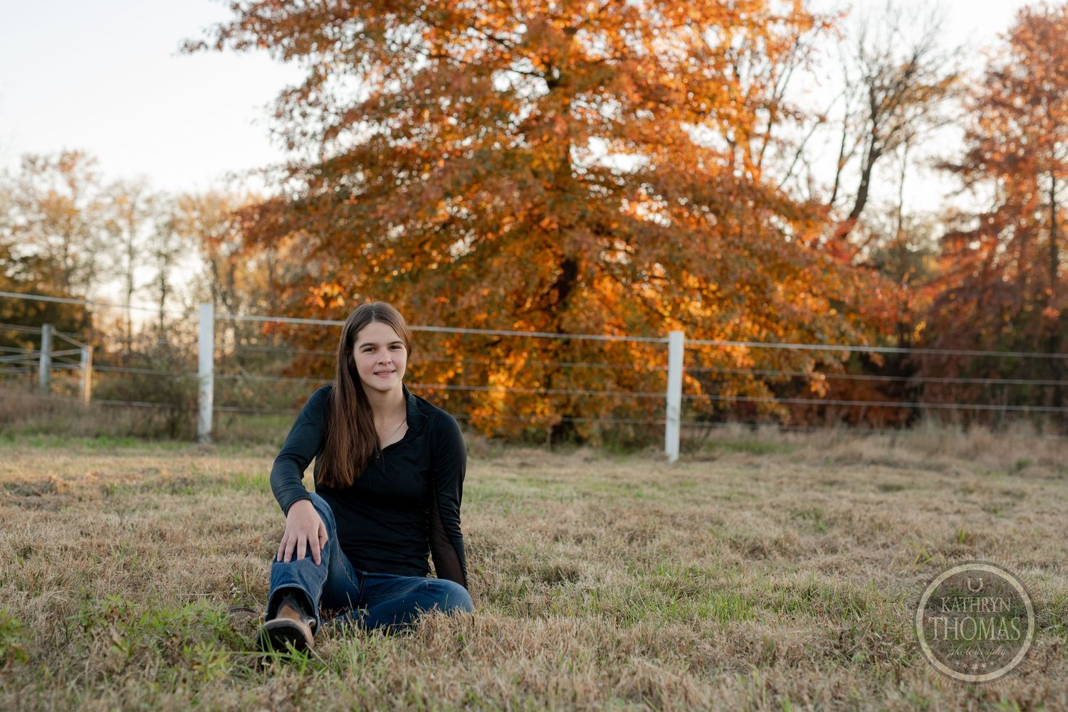 Casual sitting pose in field with fall foliage and fencing.