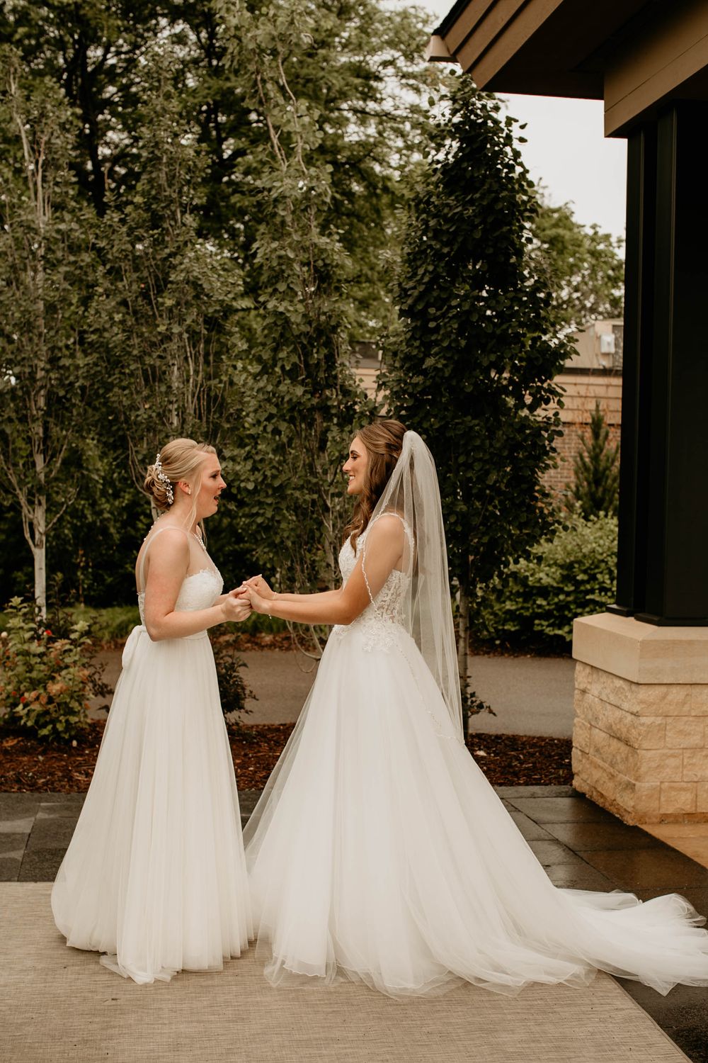 Two brides in elegant white wedding dresses hold hands and smile at each other during their first look.