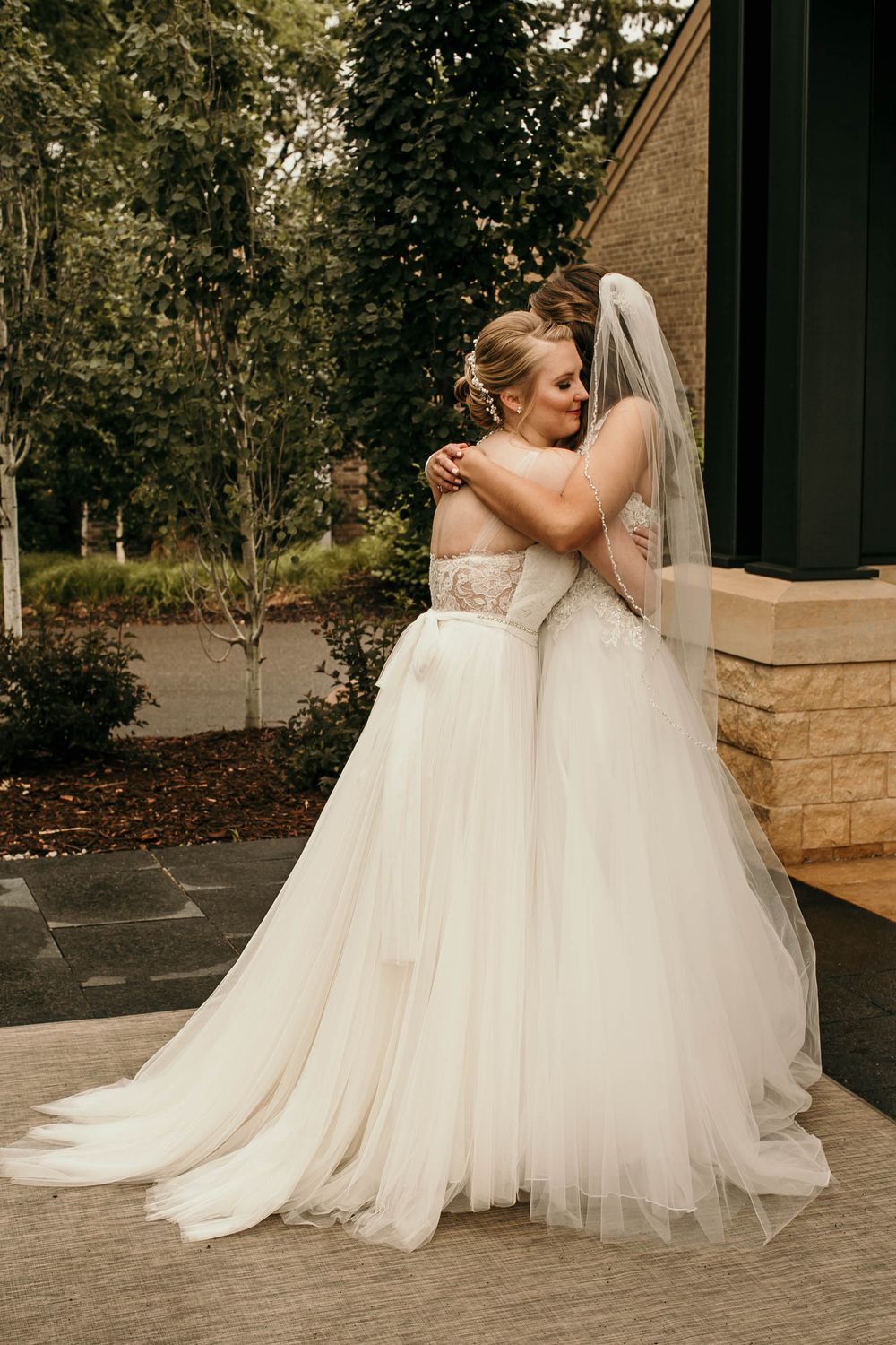 Two brides in flowing white tulle gowns share a loving embrace outside their wedding venue.