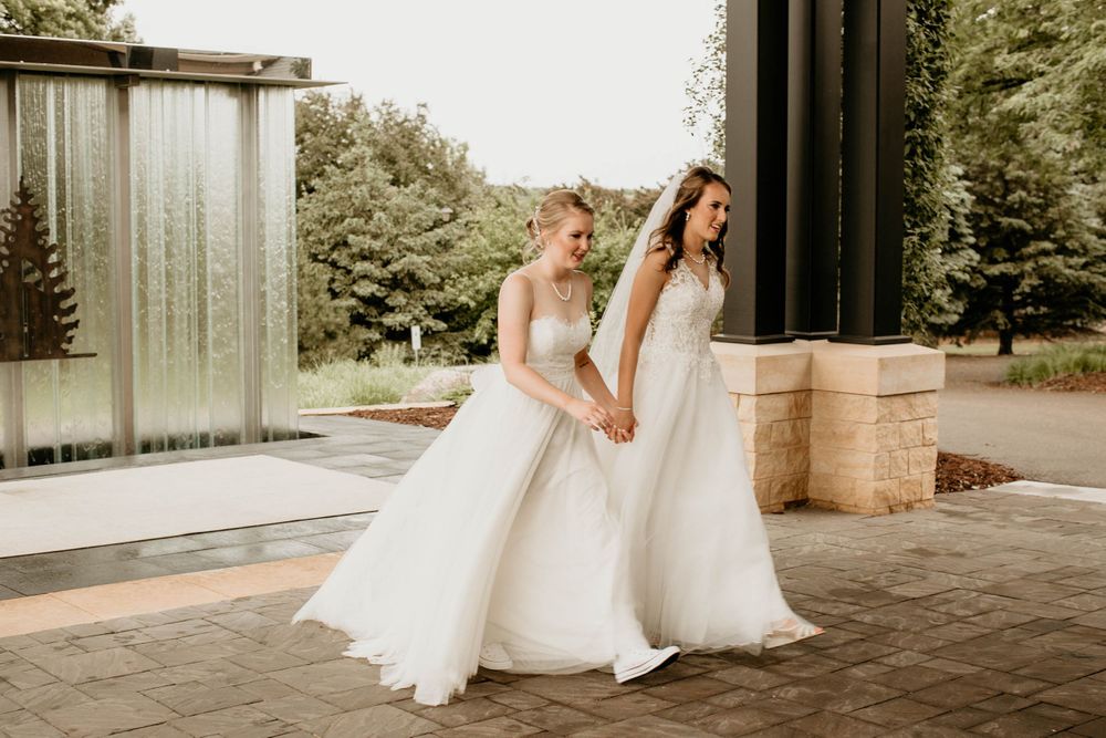 Two brides in white wedding gowns walk hand in hand past a stone wall and water feature outdoors.