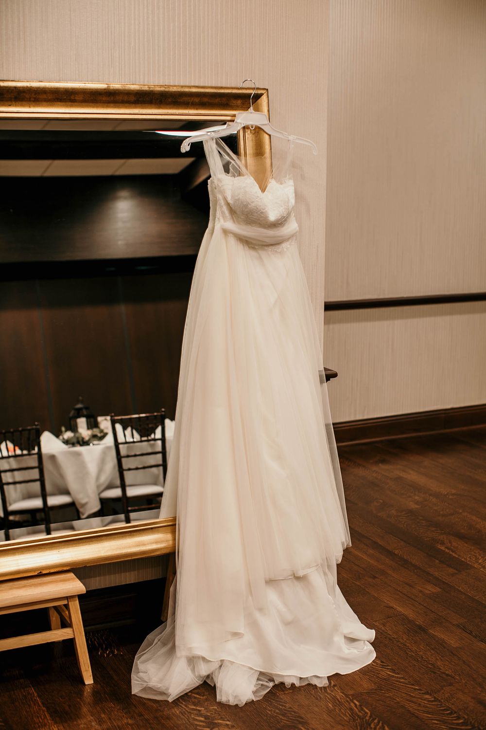 Elegant white A-line wedding dress hanging on a hanger in front of a gold-framed mirror in a bridal suite.
