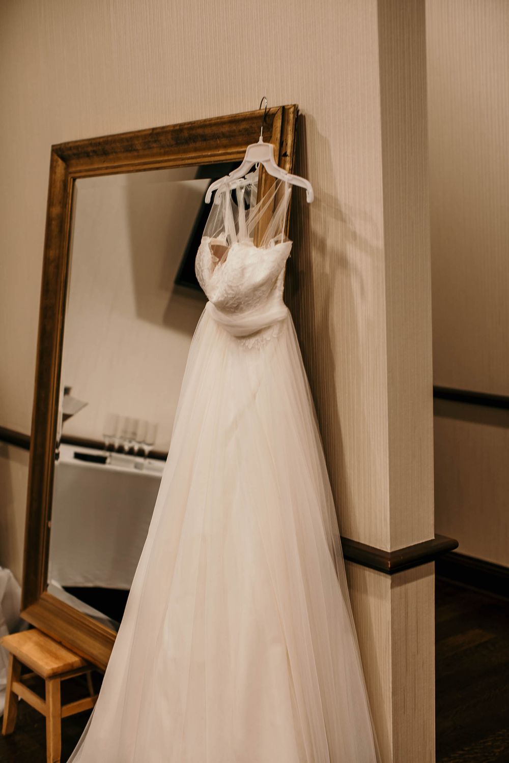 Beautiful white tulle wedding gown hanging on a white hanger beside a large ornate gold-framed mirror.