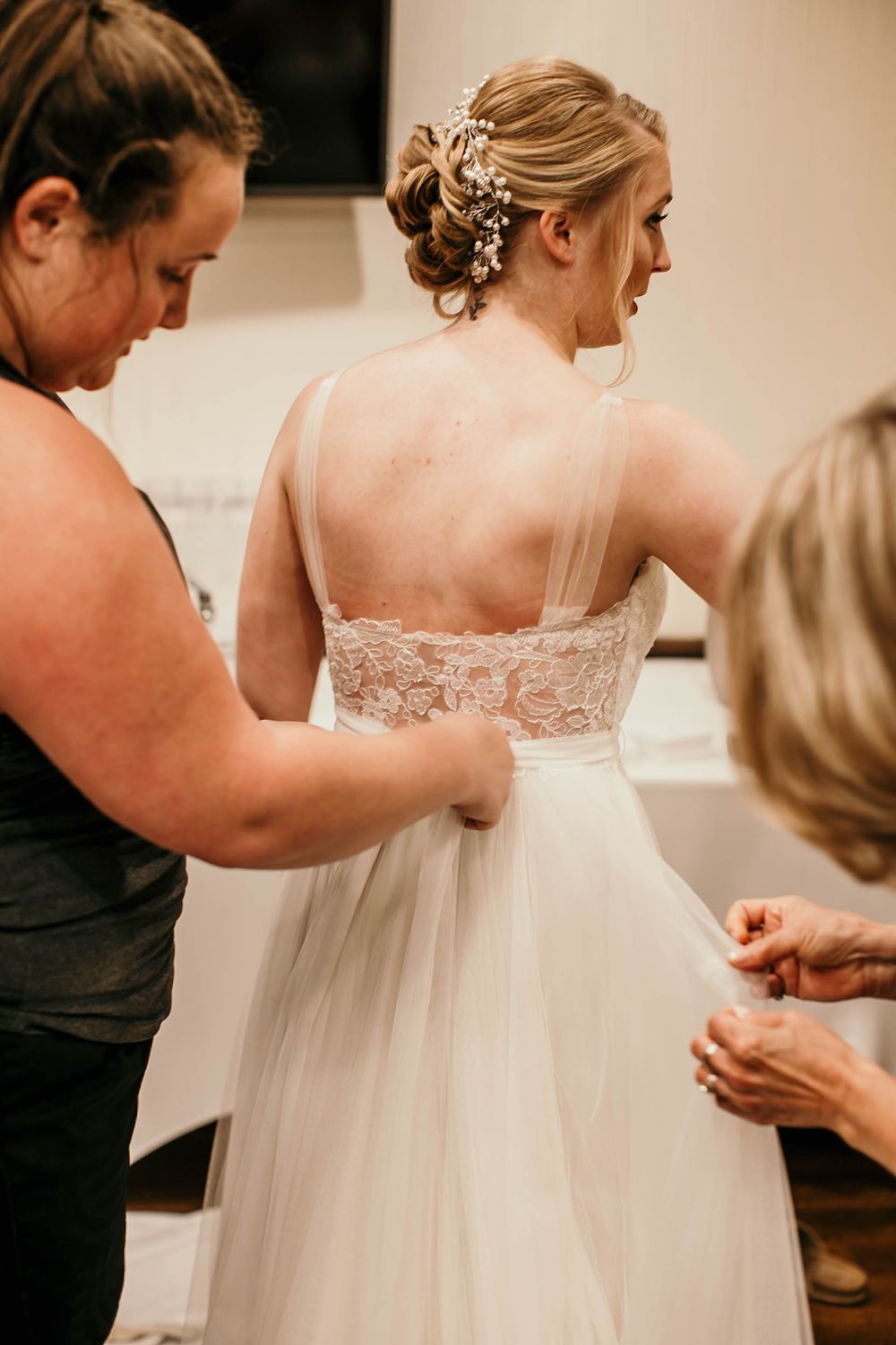 Bride with floral hair accessory getting her lace-back wedding dress fastened by two helpers before ceremony.