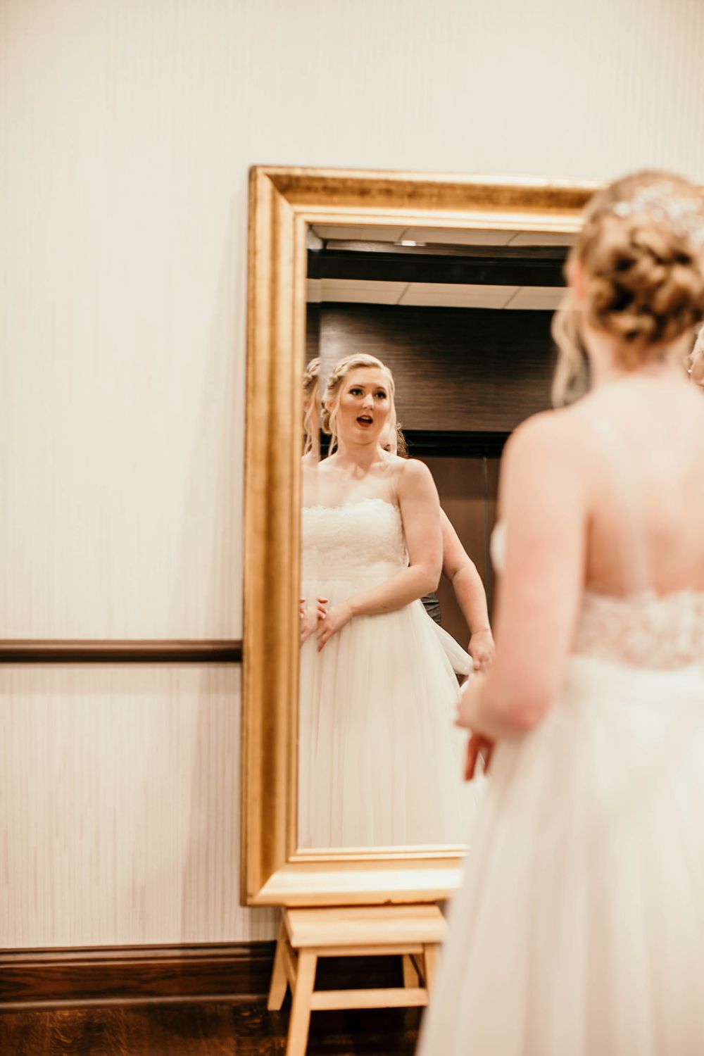 Bride in white wedding gown with updo hairstyle admiring herself in a large gold-framed dressing mirror.