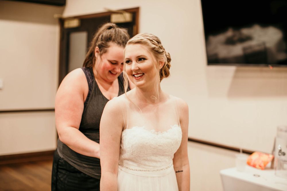 Smiling bride in strapless lace wedding dress being helped by a woman in a gray tank top before wedding.