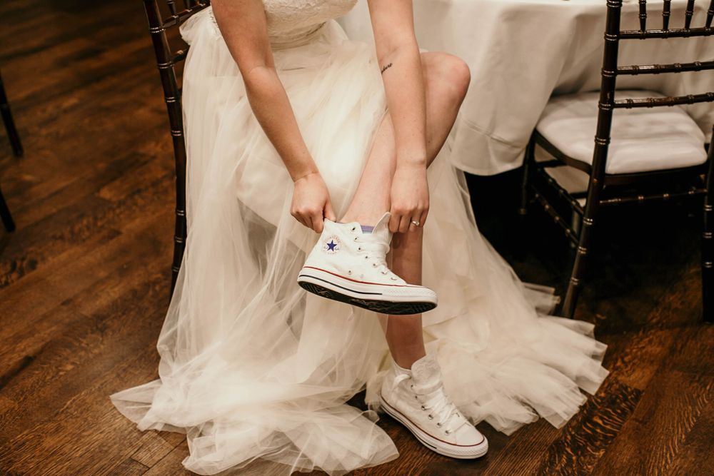 Bride in white tulle wedding gown putting on white high-top Converse sneakers while seated on wooden chair.