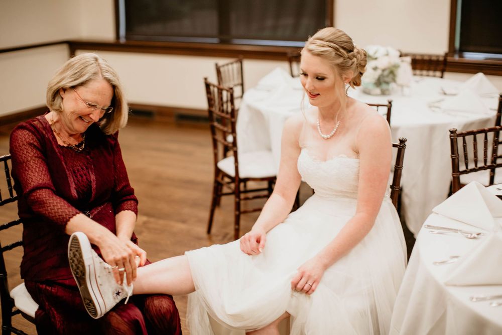 Older woman helping a smiling bride in a strapless wedding dress put on white sneakers before the ceremony.