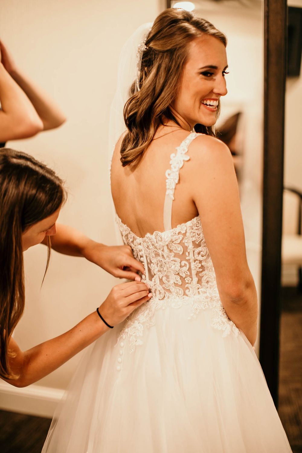 Smiling bride with brown hair getting her floral lace-back wedding dress buttoned by a bridesmaid before ceremony.