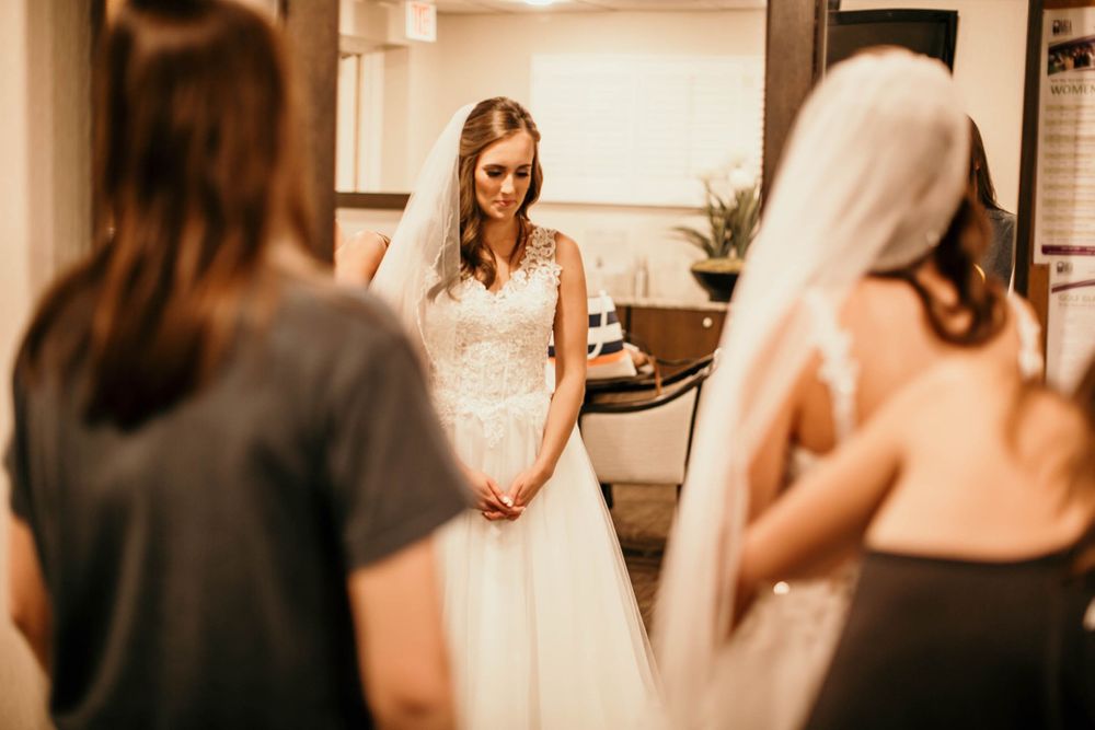 Bride in lace wedding gown stands with clasped hands as attendants help her prepare for her special day.