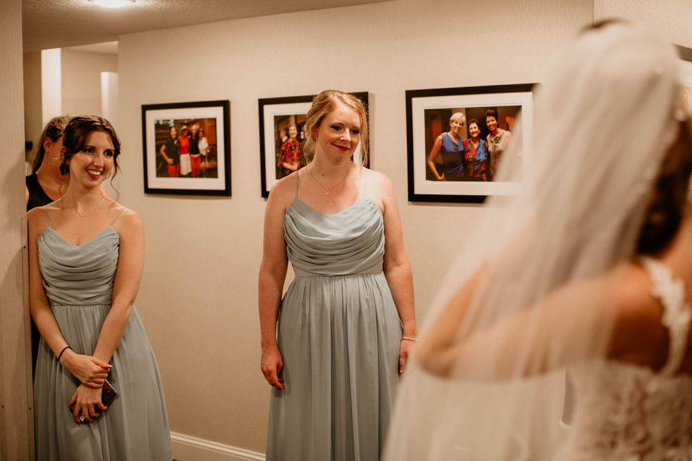 Two bridesmaids in sage green gowns smile with joy upon seeing the bride in her wedding dress.