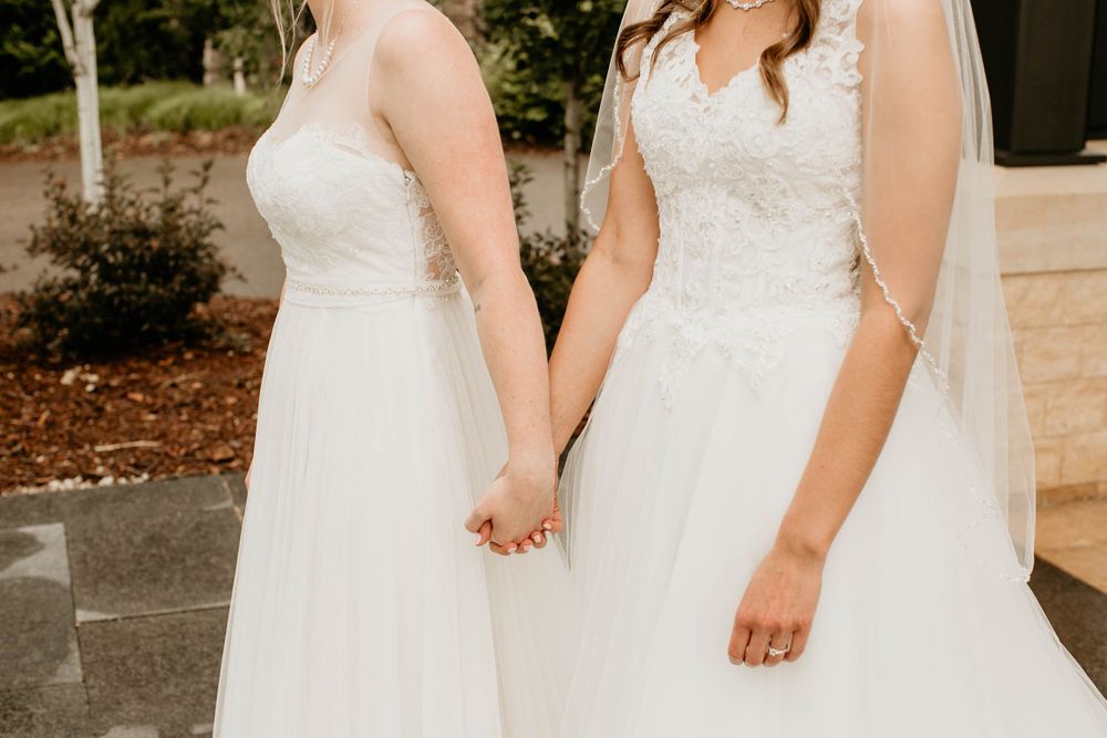 Two brides in flowing white wedding dresses hold hands in a close romantic moment on their wedding day.