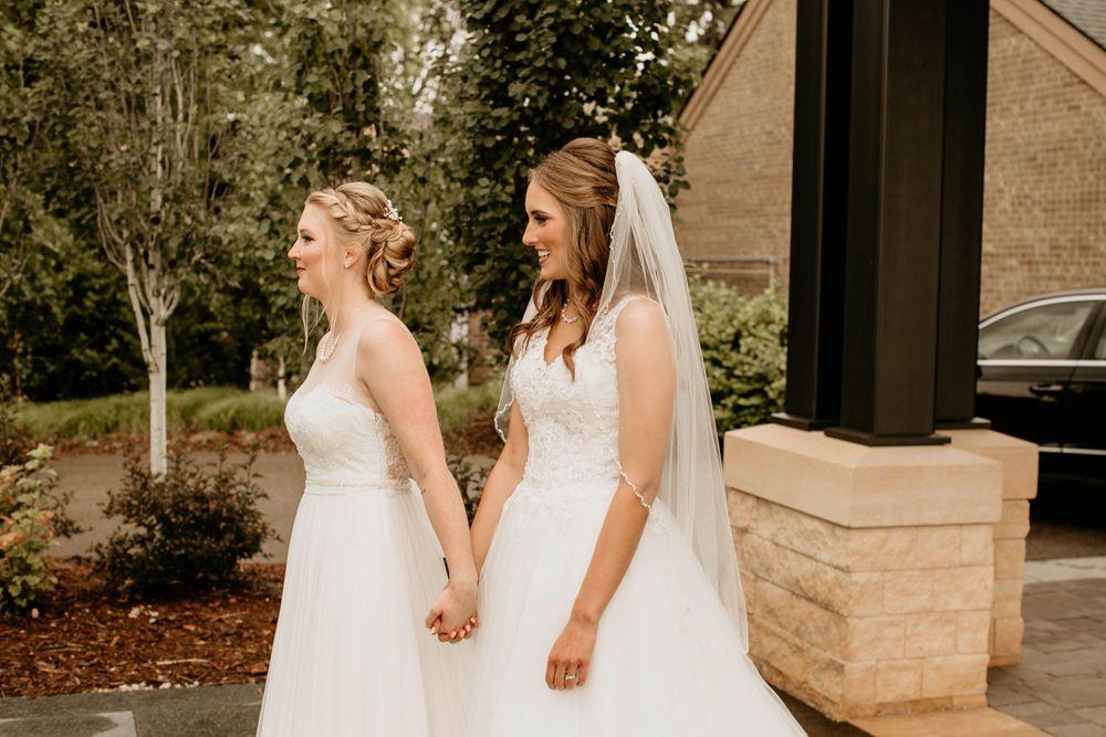 Two brides in white wedding gowns hold hands and share a tender moment outdoors on their wedding day.