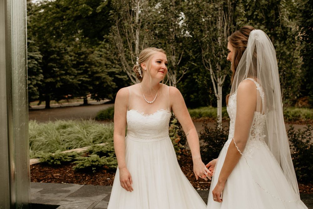 Two brides in elegant white gowns laugh and hold hands together during their outdoor wedding first look.