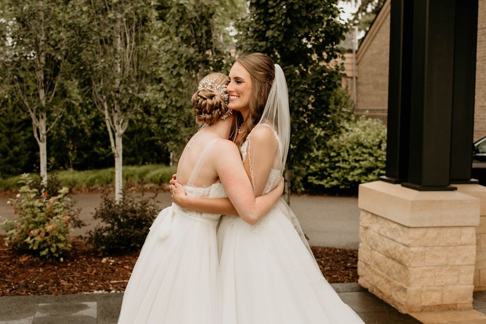 Two brides in white wedding gowns share a warm embrace during an emotional outdoor first look moment.