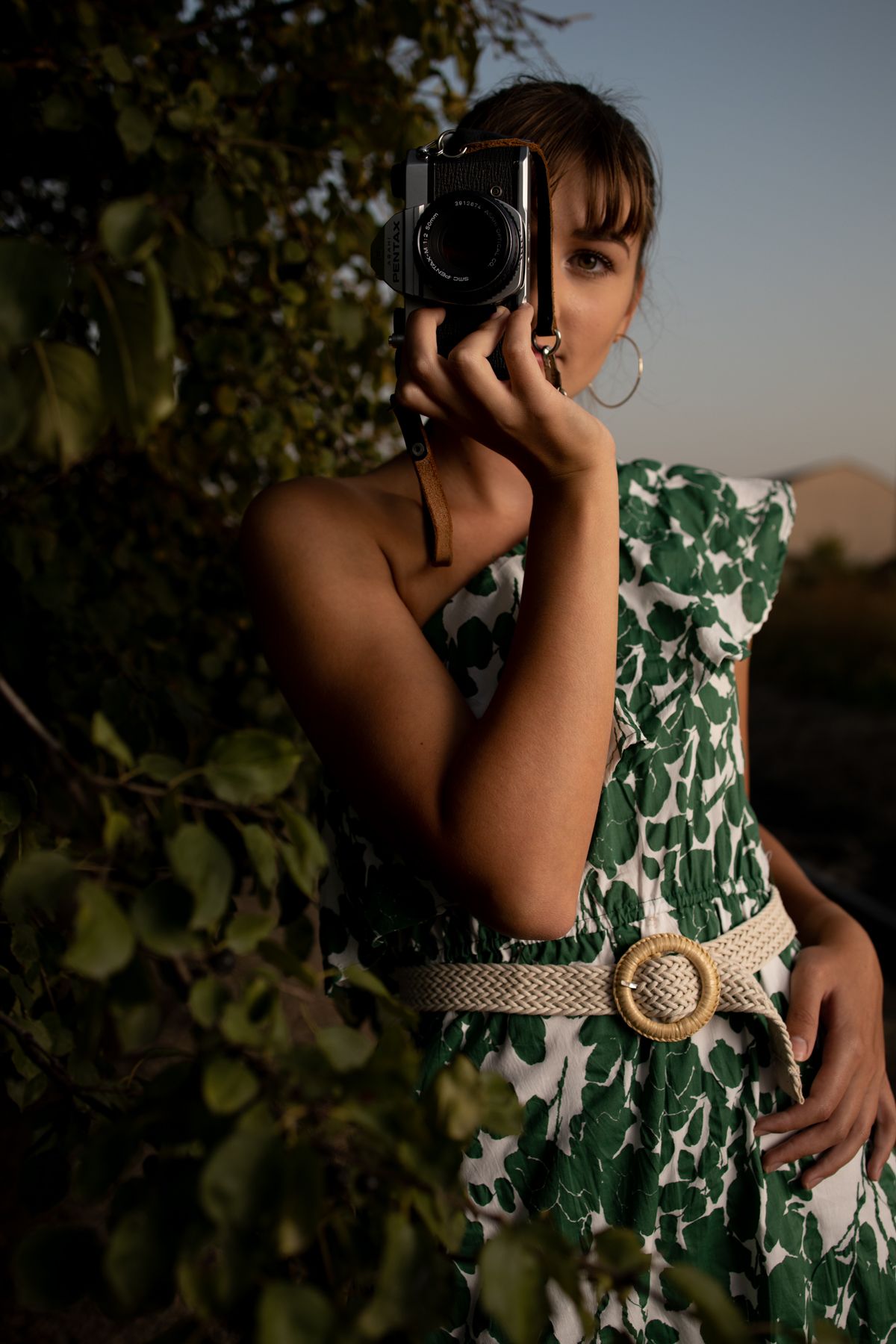 Photographer in green and white floral dress holds vintage camera against sunset-lit foliage backdrop. photographer Gerardo Dominguez