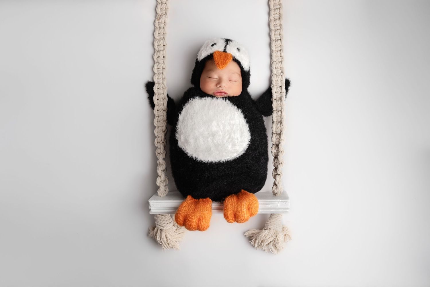 A newborn dressed in a penguin costume sleeps on a small knitted rope swing against a white background.