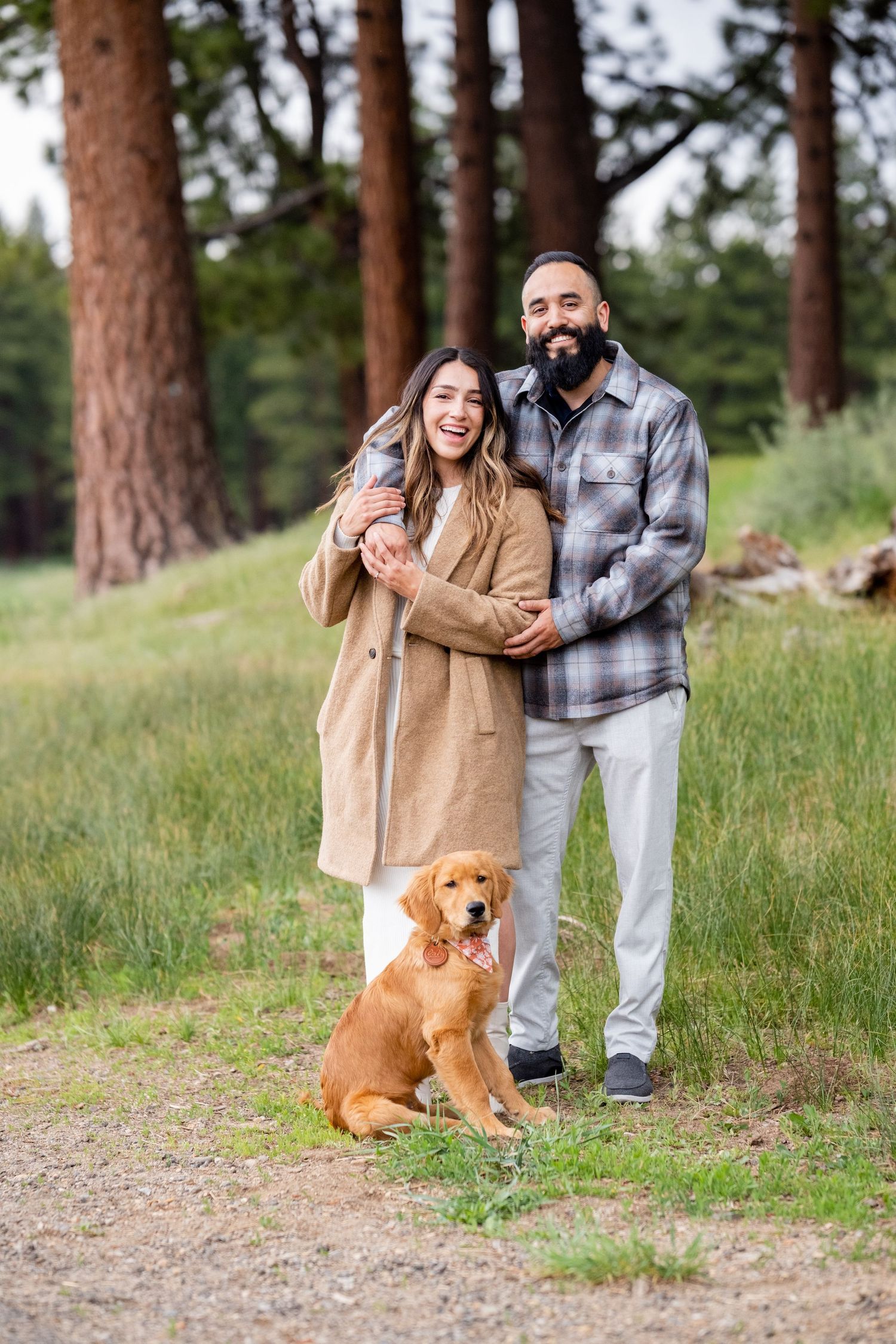 A couple embraces on a forest trail with their golden retriever sitting obediently beside them on a sunny day.