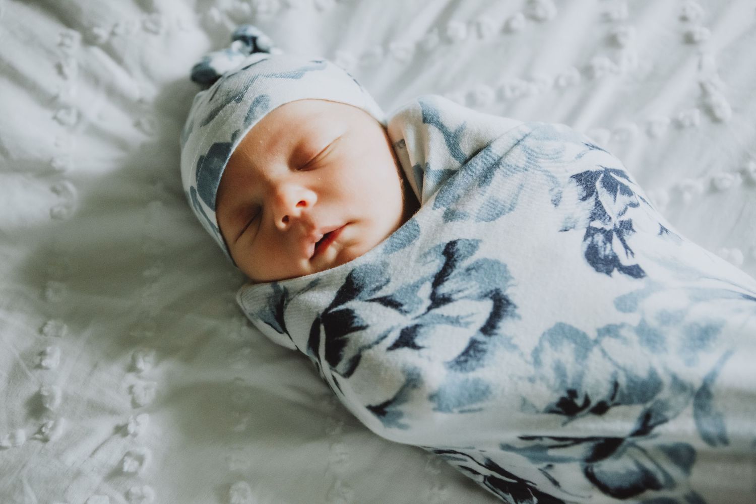 A peaceful newborn sleeping in a blue floral swaddle on white textured bedding.