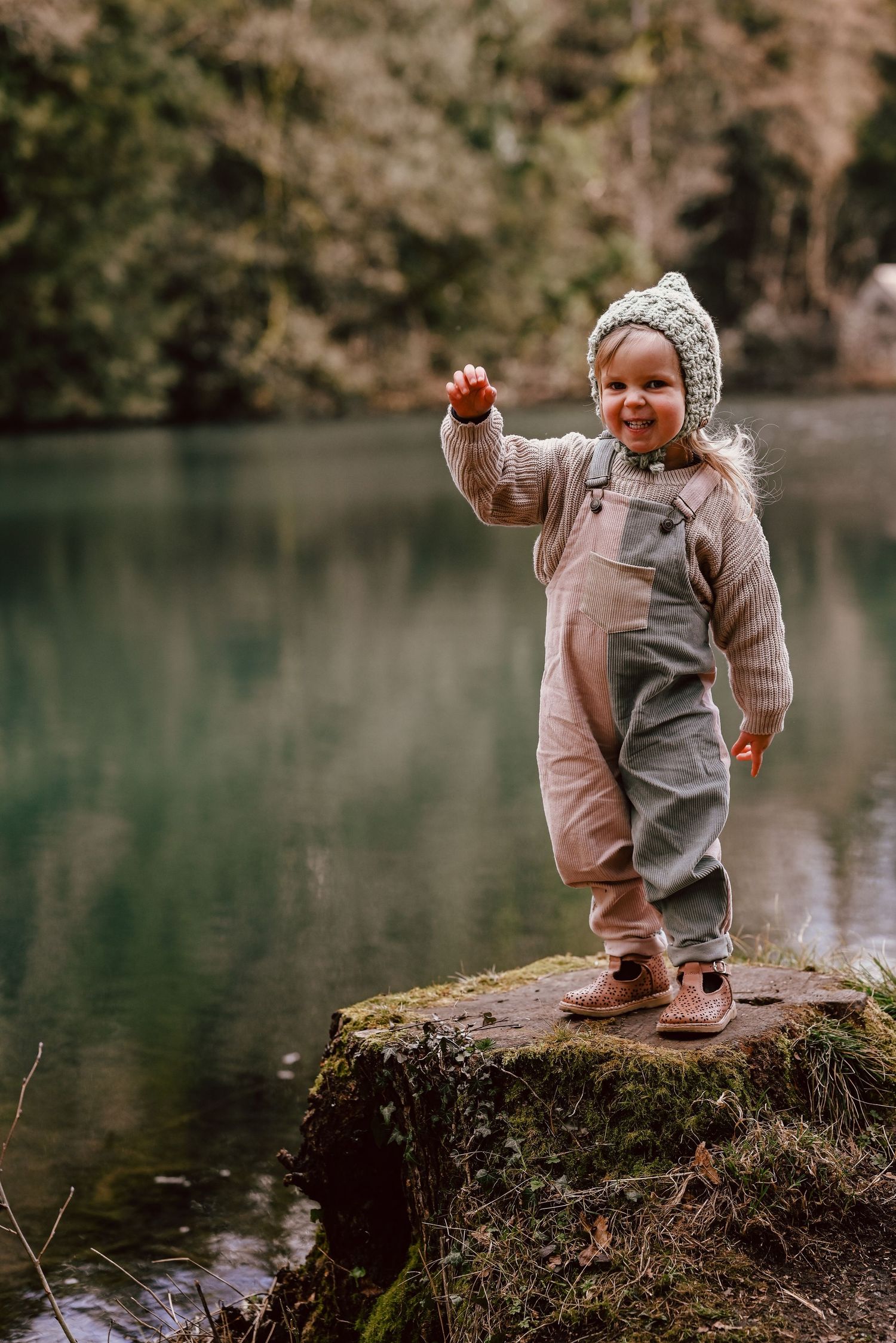 A toddler in cozy winter wear stands on a mossy rock by a serene lake while holding something up in excitement.
