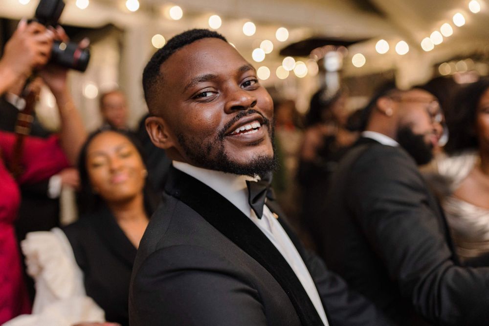 Elegantly dressed guest in a black tuxedo and bowtie smiles warmly at a wedding reception with twinkling lights.