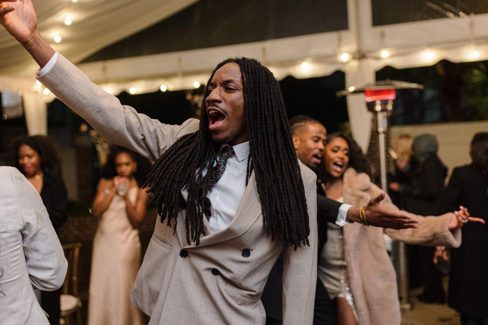 Guest in light colored suit dancing with raised arms at wedding celebration.