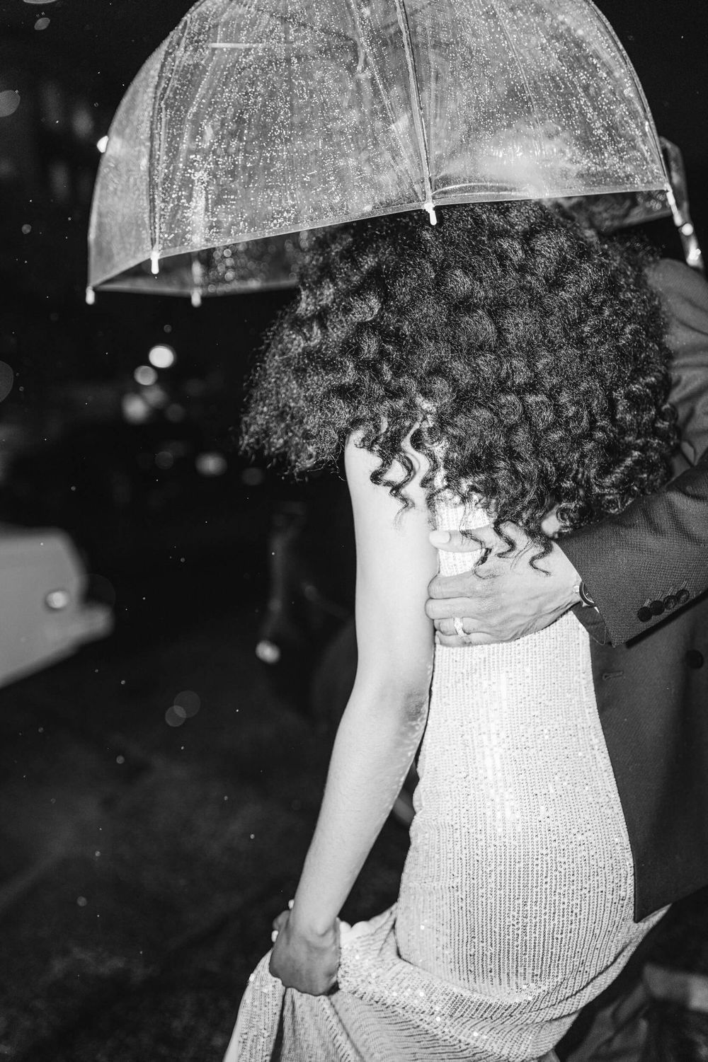 Person holding clear umbrella in rain wearing sparkly dress at night event.