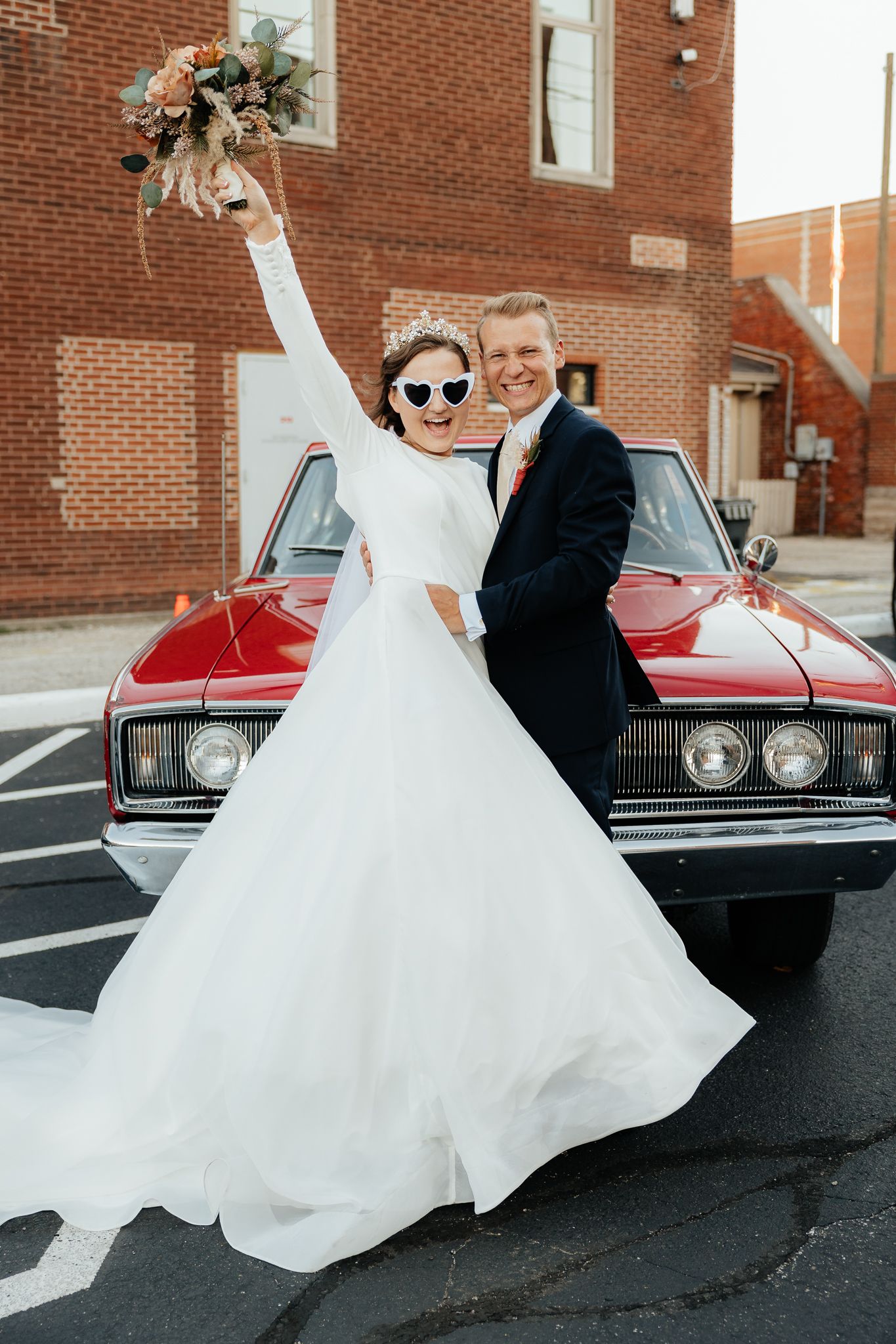 Newlyweds posing playfully by a vintage red car outside a brick building.