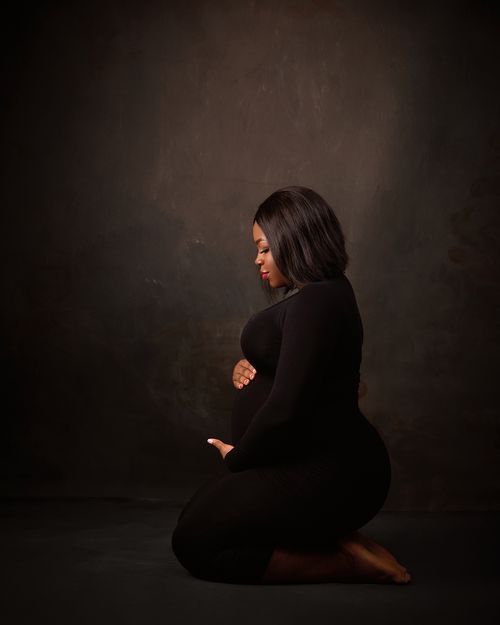 Low-key maternity portrait of woman in black dress kneeling against dark background.