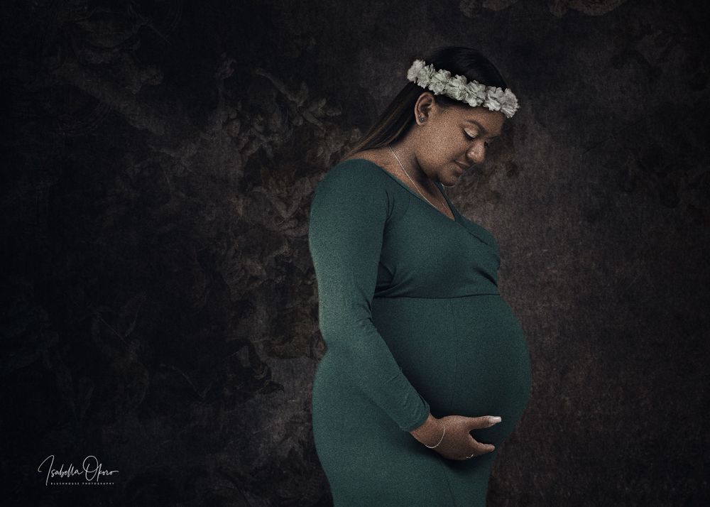 Artistic maternity photo of pregnant woman in green dress with floral crown against dark backdrop.