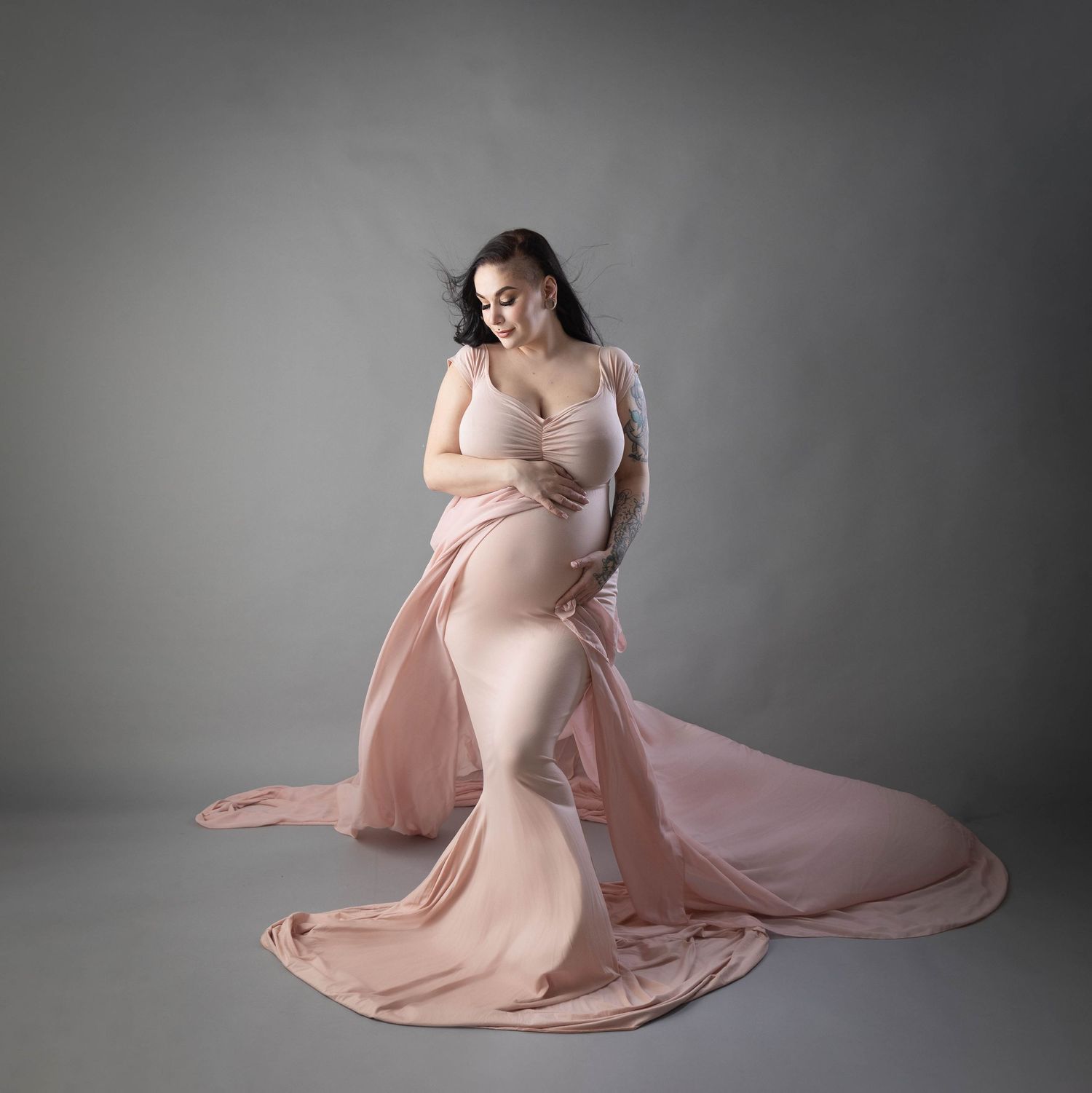 Dreamy maternity photo in blush pink flowing gown against neutral backdrop.
