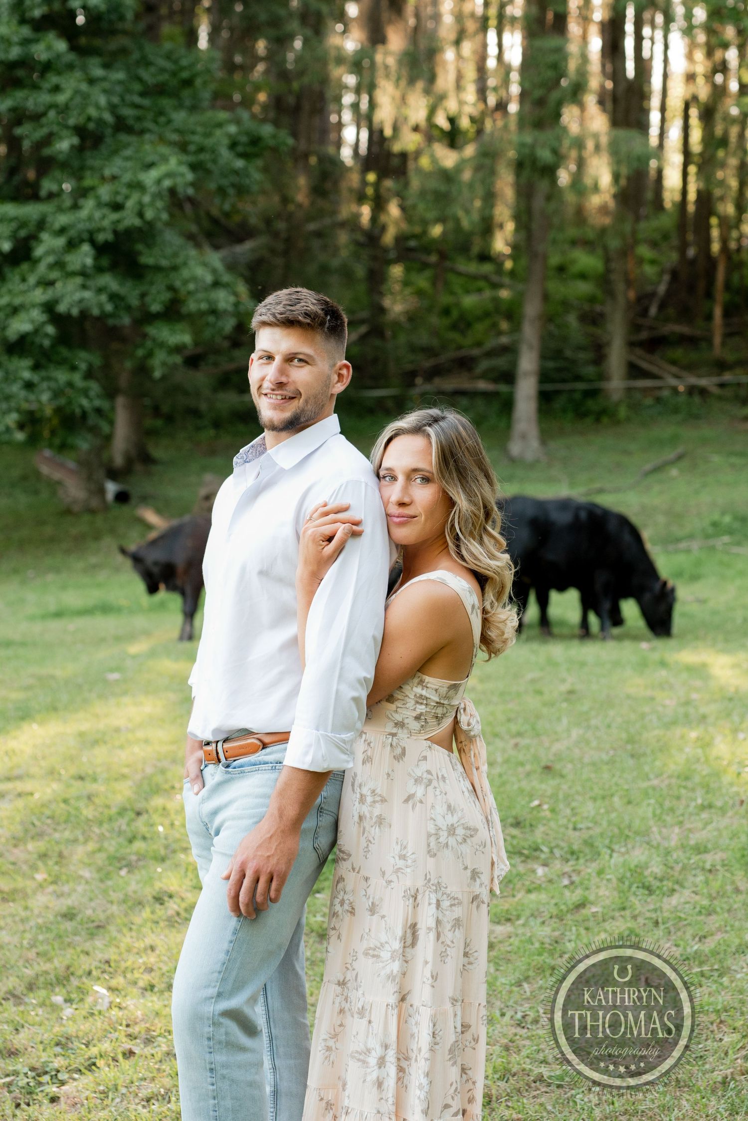 A couple poses in front of grazing cattle on a grassy hillside near a forest.