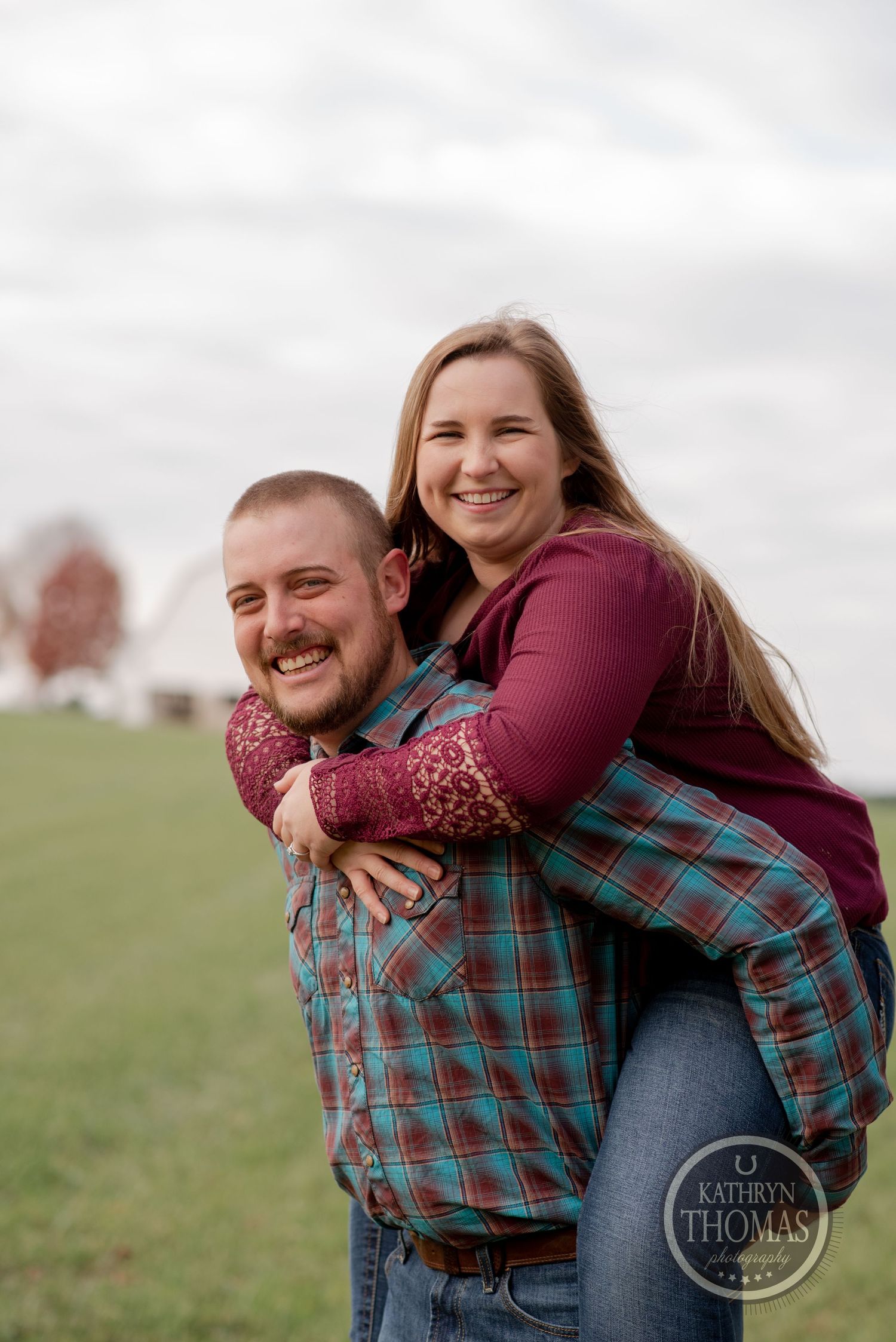 A couple shares a playful piggyback ride moment in a grassy field on an overcast day.