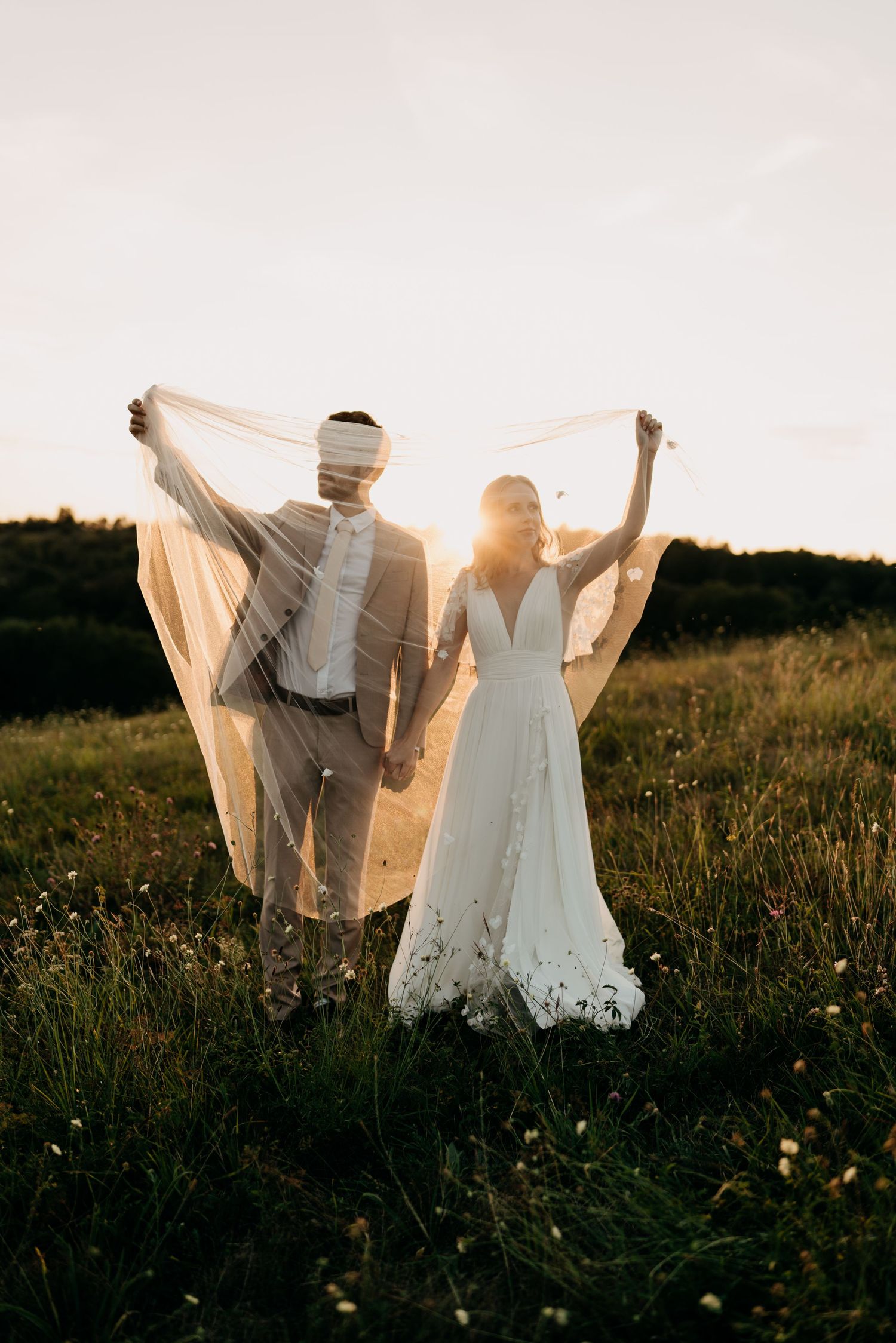 Bride and groom hold flowing veil in a sunlit meadow during golden hour wedding shoot.