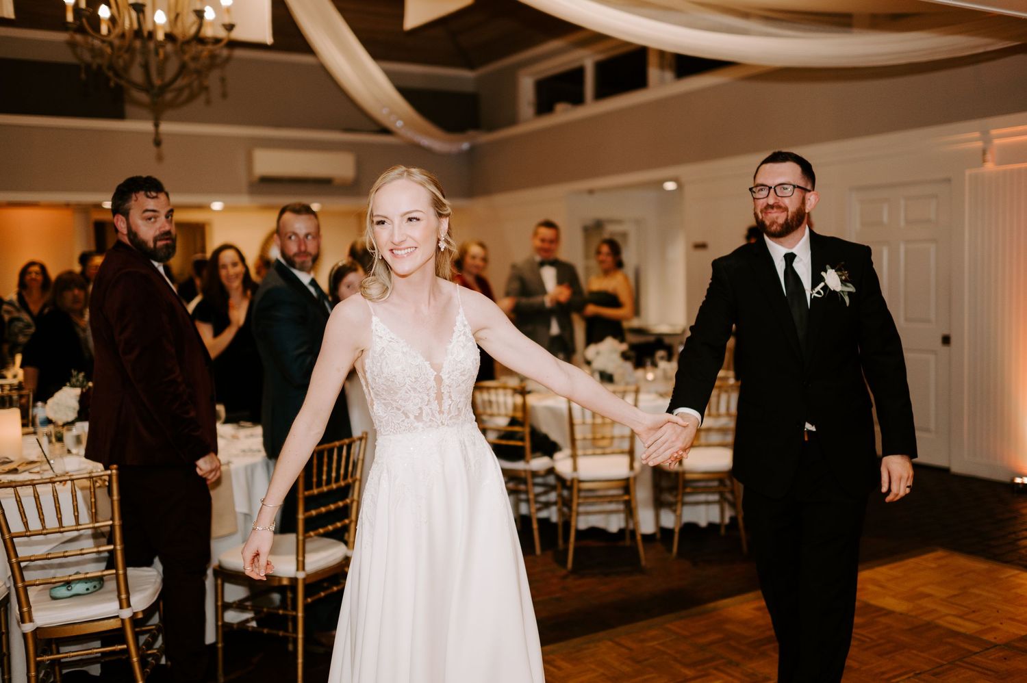 Wedding reception features elegant decor with white draping and chandelier lighting.