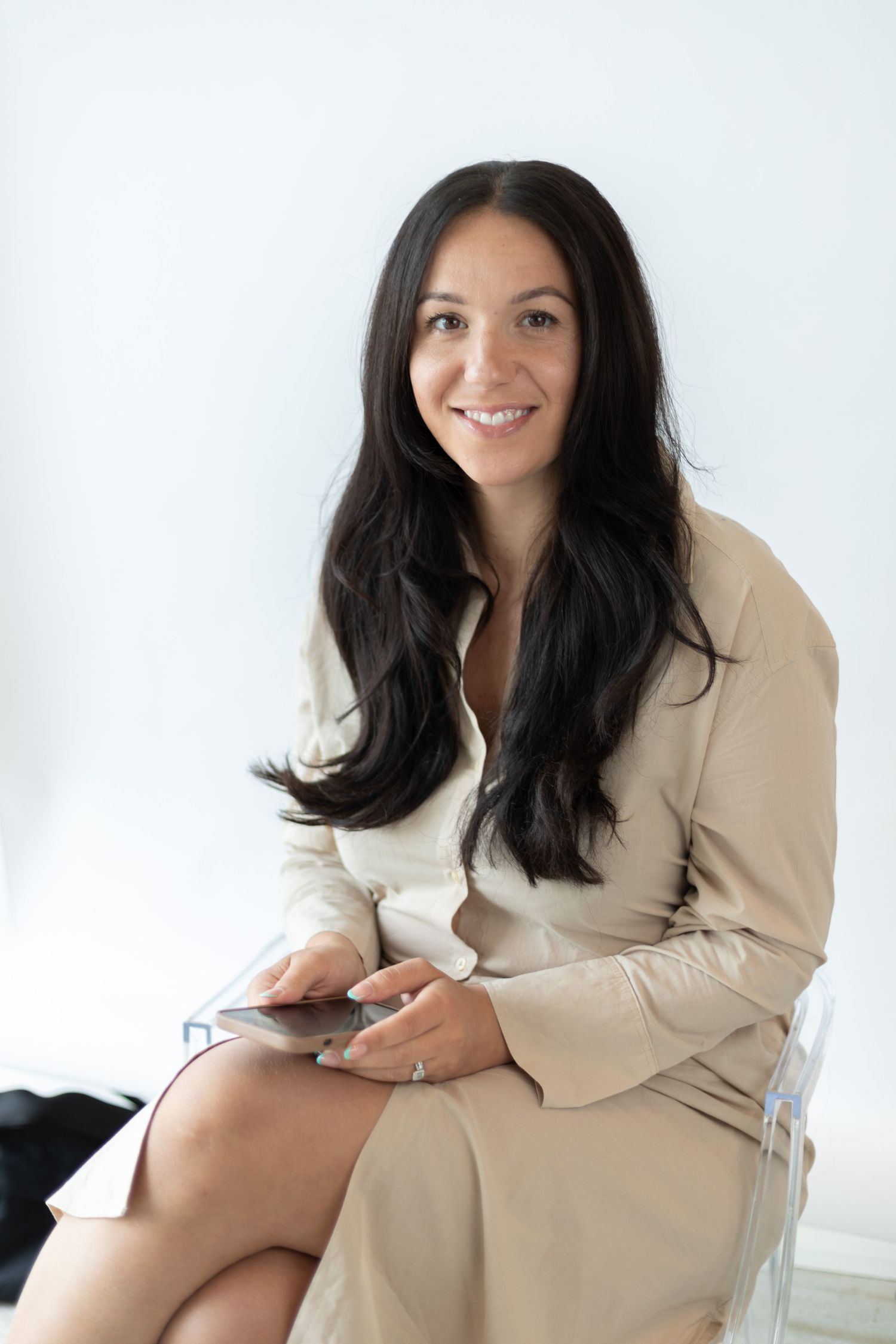 Woman in beige blazer sitting on clear chair while holding phone.