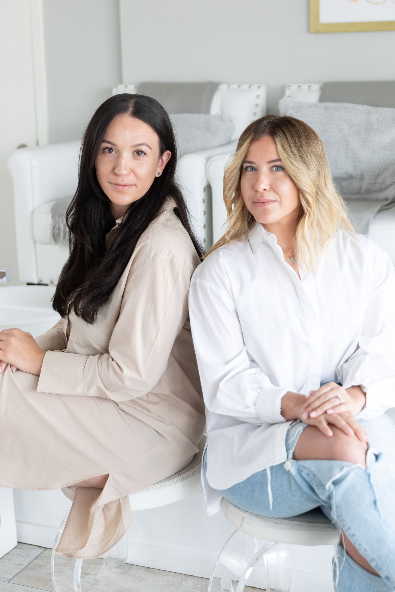 Two women sitting together on white furniture wearing casual business attire.